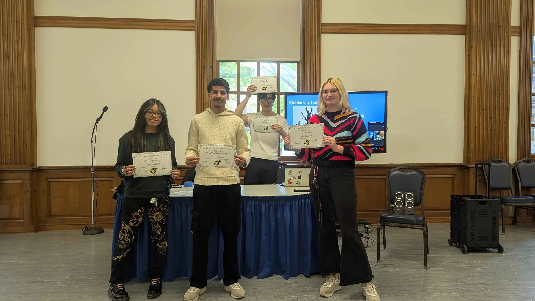 Four students holding certificates at the front of a hall with wood paneling